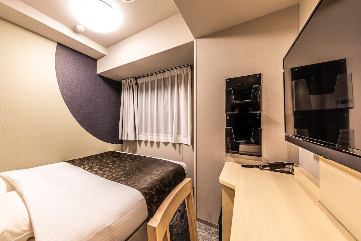 Une chambre à coucher de taille réduite, comprenant un lit, un bureau, une chaise, un téléviseur et une fenêtre avec des rideaux.