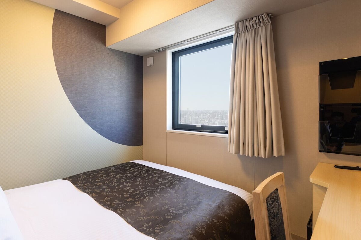 Une chambre d’hôtel avec un lit, un bureau, une chaise, une fenêtre avec des rideaux et une vue sur la ville.