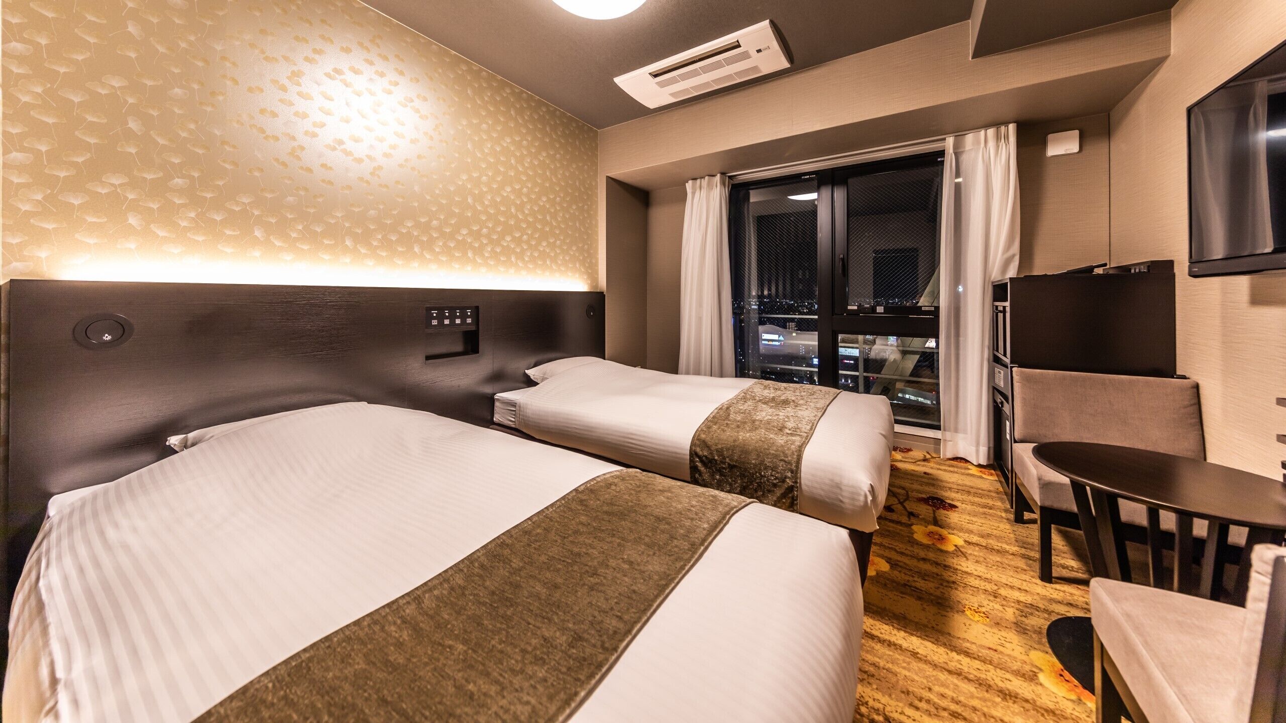 Ein Hotelzimmer mit zwei Betten, einem Schreibtisch, einem Sessel, einem Fernseher und einem Fenster mit Blick auf die Stadt.