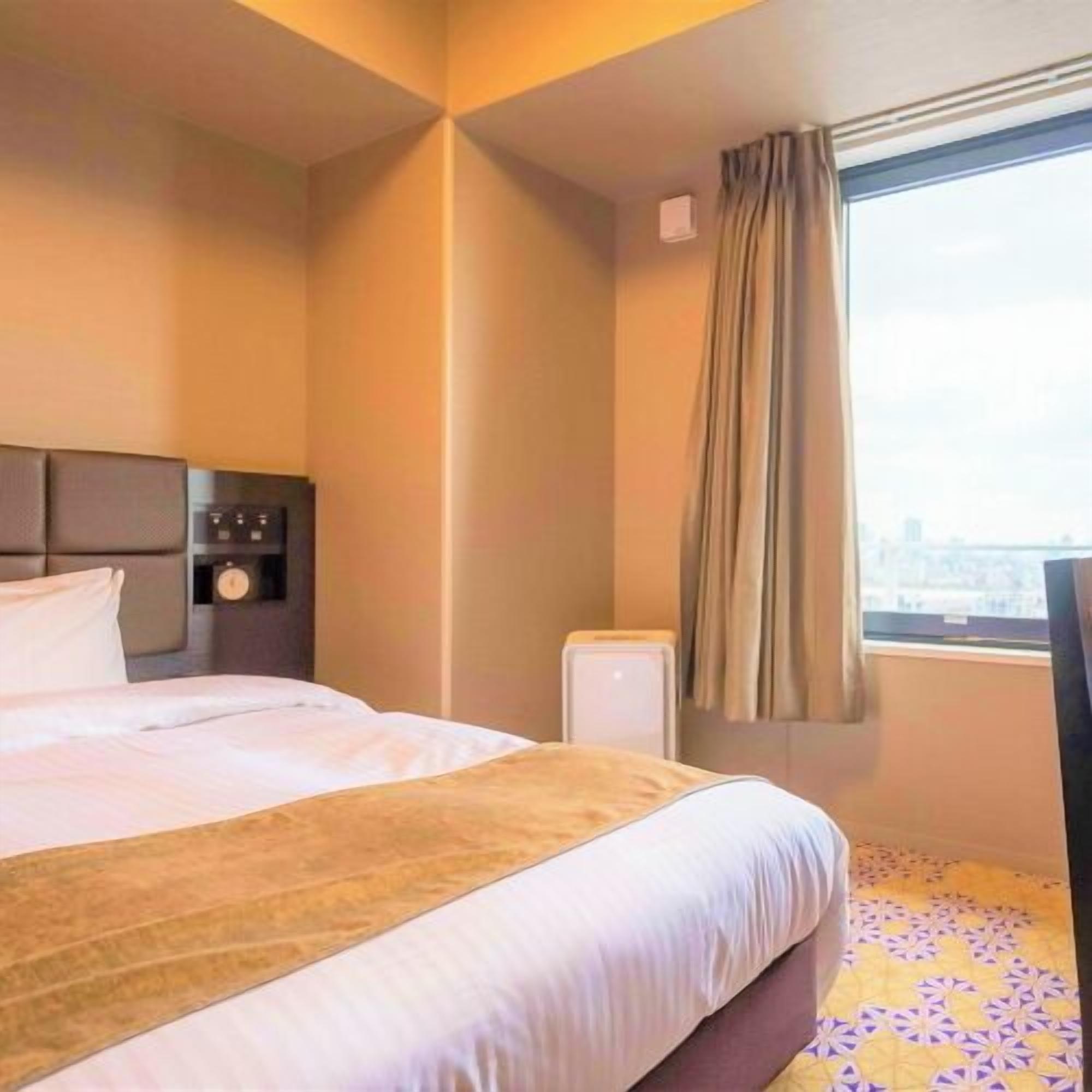 Ein Hotelzimmer mit Bett, Nachttisch, einer Uhr, einem Fenster mit Vorhängen und Blick auf die Stadt.
