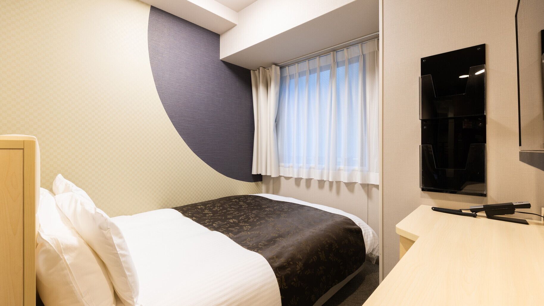 Une chambre d’hôtel équipée d’un lit, d’un bureau, d’une fenêtre avec des rideaux et d’un téléviseur fixé au mur.