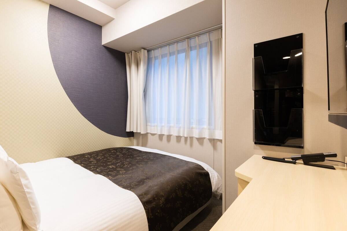 Une chambre d’hôtel équipée d’un lit, d’un bureau, d’une fenêtre avec des rideaux et d’un téléviseur fixé au mur.