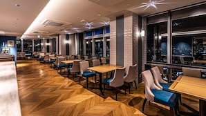 Een modern restaurant met houten visgraatvloer, meerdere tafels en stoelen, en grote ramen met uitzicht over de stad.