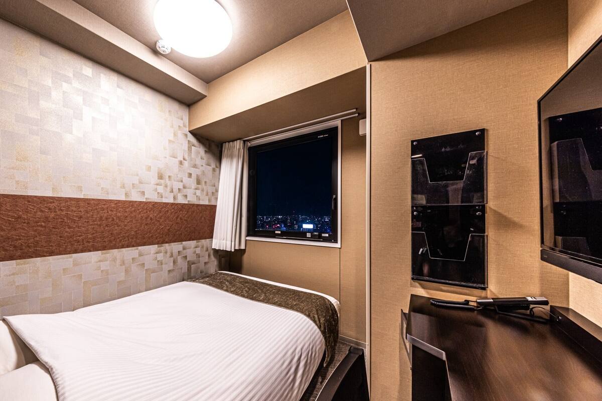 Une chambre d’hôtel avec un lit, une fenêtre donnant sur la ville, un miroir et un téléviseur fixé au mur.