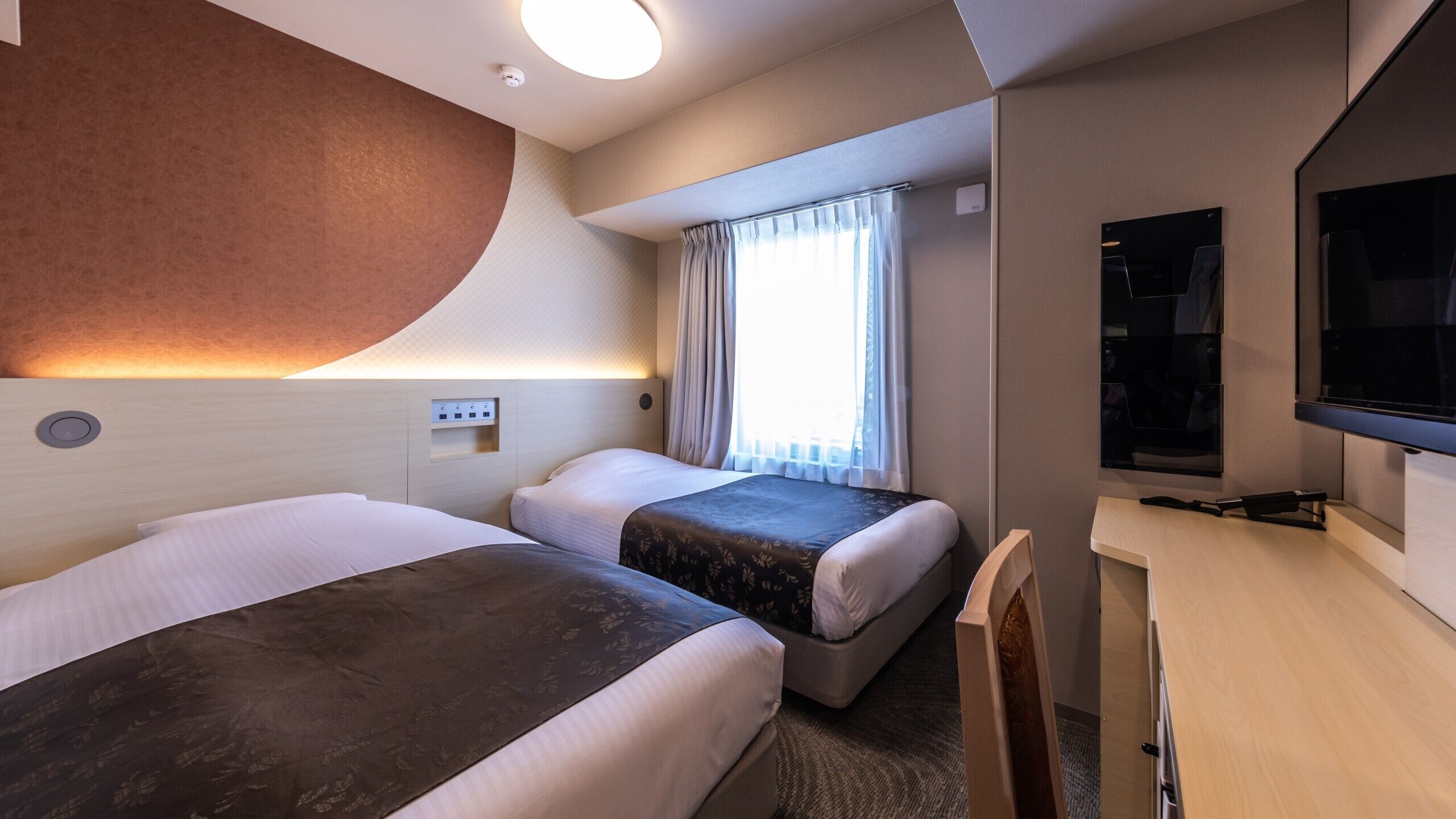 Ein Hotelzimmer mit zwei Betten, einem Schreibtisch, einem Fernseher und einem Fenster mit Vorhängen.