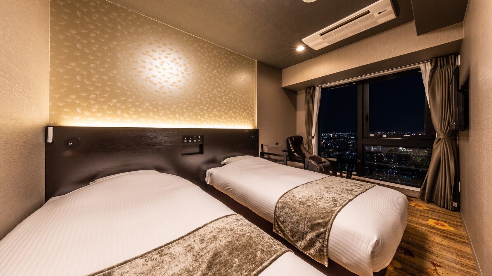 Une chambre d’hôtel avec deux lits, une table de chevet, un bureau, une chaise, une fenêtre donnant sur la ville et un appareil de climatisation.