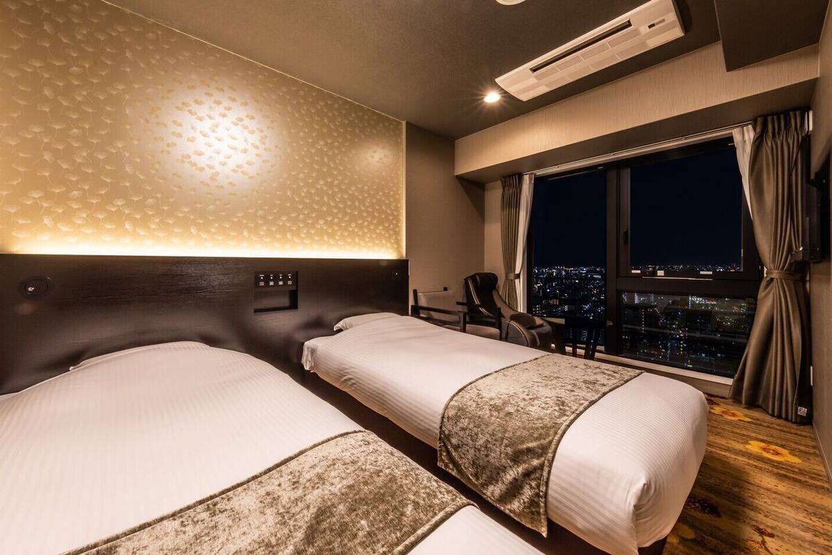 Une chambre d’hôtel avec deux lits, une table de chevet, un bureau, une chaise, une fenêtre donnant sur la ville et un appareil de climatisation.