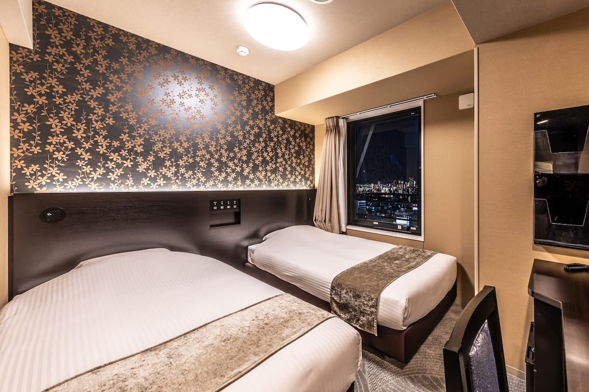 Une chambre d’hôtel avec deux lits, une grande fenêtre donnant sur la ville illuminée la nuit, un minibar et une télévision.