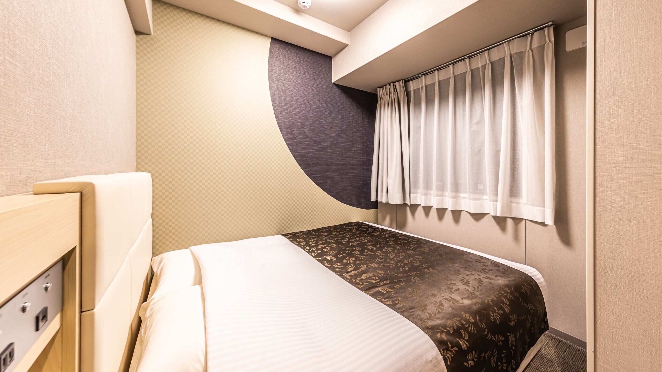 Ein kleines Hotelzimmer mit Bett, Bedienfeld, Fenster mit Vorhängen und einer wandgebogenen Gestaltung.