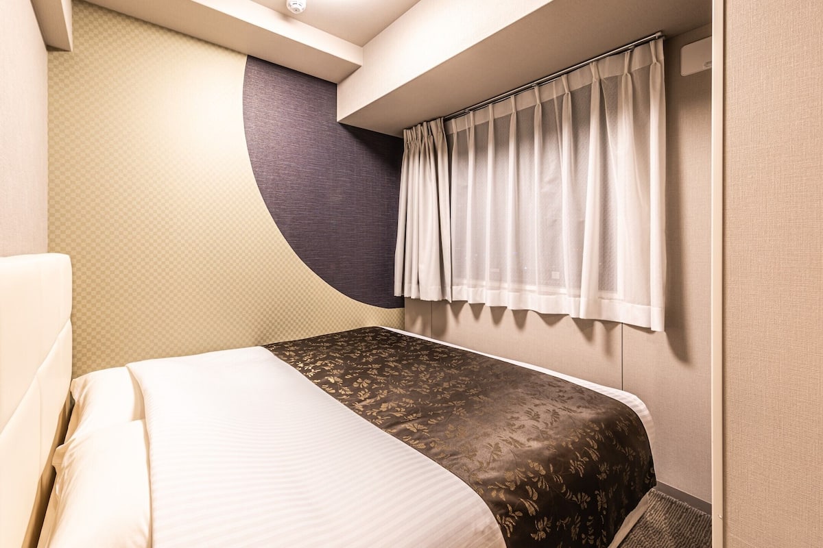 Une chambre d’hôtel compacte comprenant un lit, un panneau de commande, une fenêtre avec des rideaux et un mur au design incurvé.