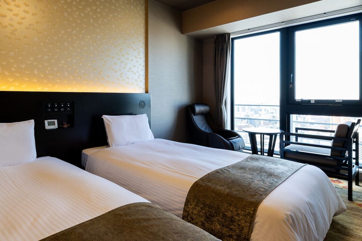 Une chambre d’hôtel avec deux lits, un panneau de commande, un fauteuil de massage et une vue sur la ville.