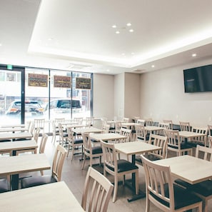 A restaurant interior with multiple tables and chairs, a large window with a view of parked cars, and a flat-screen TV mounted on the wall. - Kuretake Inn Osaka Midousuji Hommachi (Osaka)
