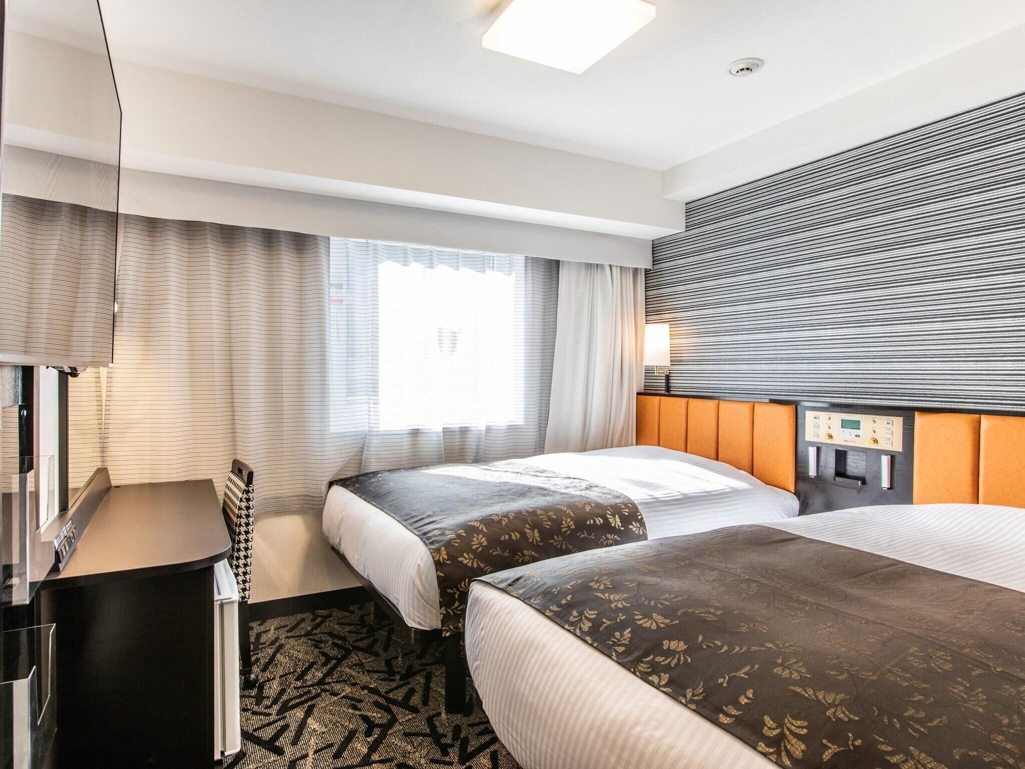 Ein Hotelzimmer mit zwei Betten, einem Schreibtisch, einem Fenster mit Vorhängen und einer Bedienfeld an der Wand.