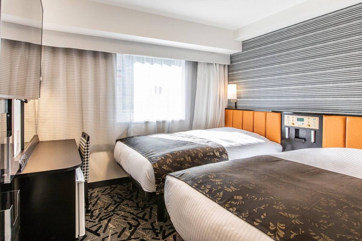 A hotel room with two beds, a desk, a window with curtains, and a control panel on the wall.