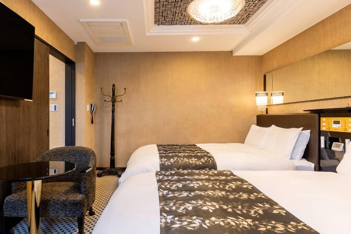 A hotel room with a bed, a desk, a chair, a TV, a coat rack, and a ceiling light.
