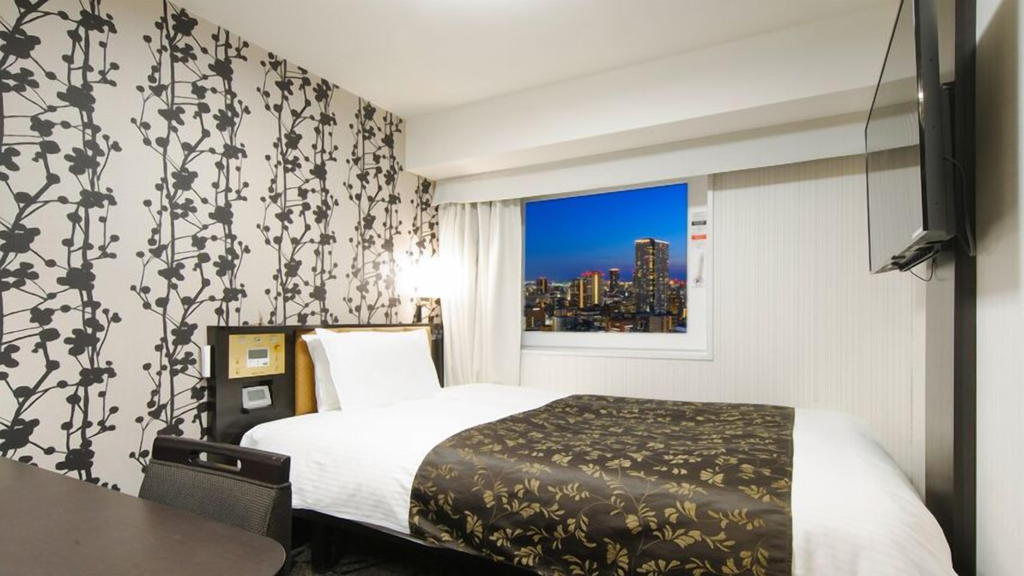 Ein Hotelzimmer mit Bett, Schreibtisch, Stuhl, Nachttisch, Fernseher und einem Fenster mit Blick auf die Stadt.