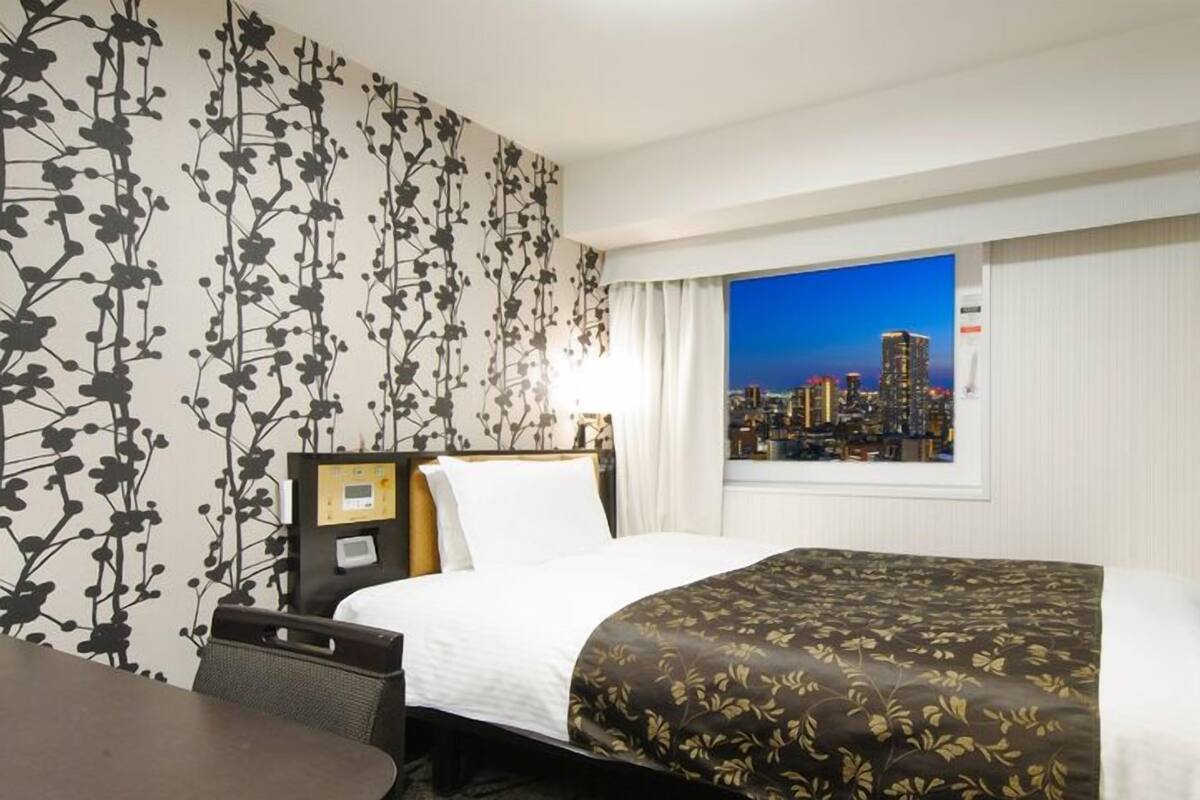 A hotel room with a bed, a desk, a chair, a nightstand, a TV, and a window with a cityscape view.
