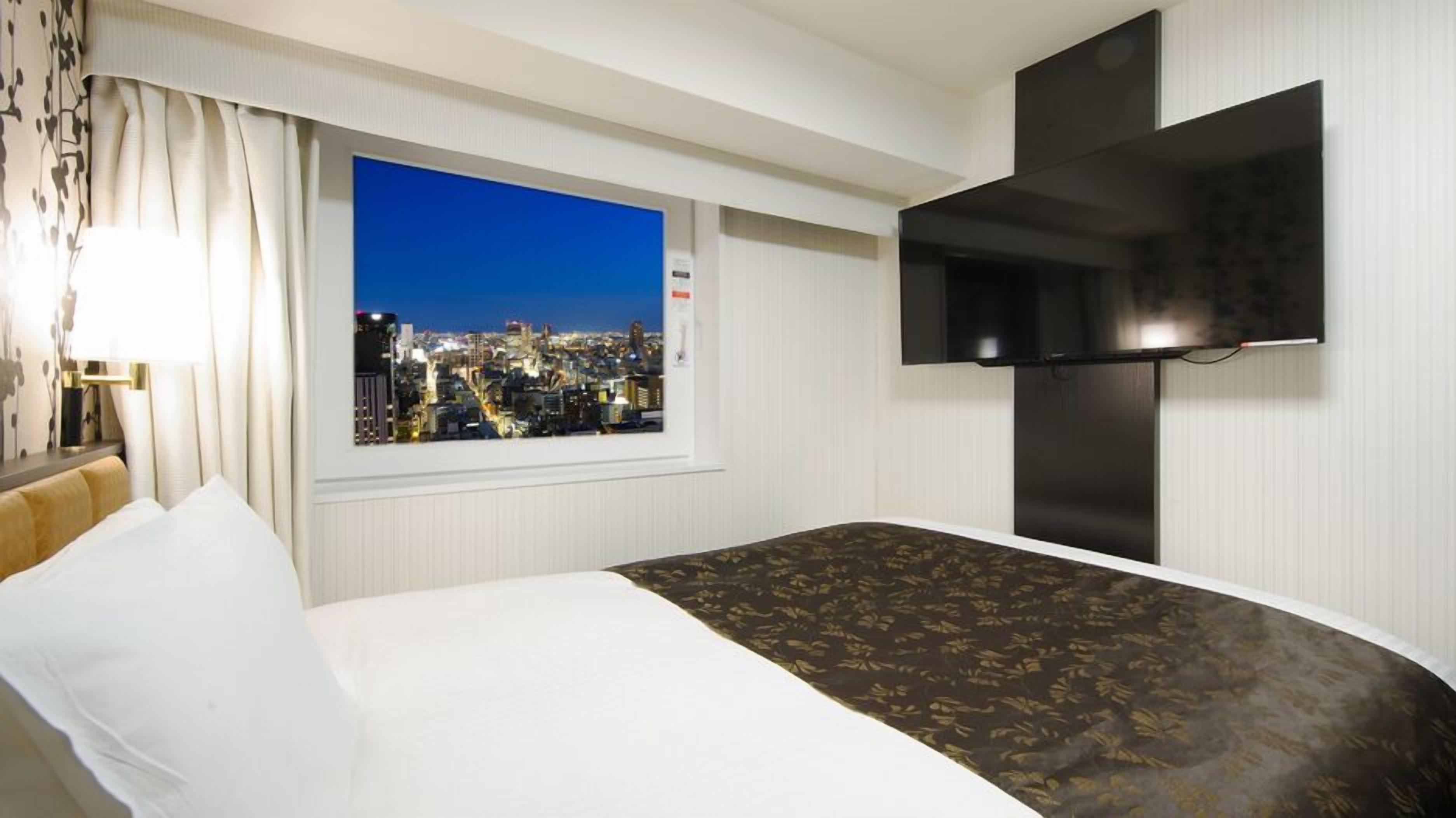 Ein Hotelzimmer mit Bett, Fenster mit Blick auf die Stadt, einem an der Wand befestigten Flachbildfernseher und einem Nachttisch mit Lampe.