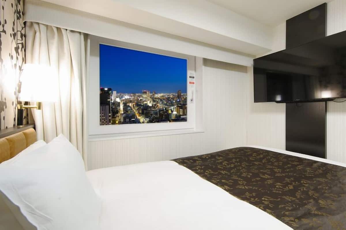 A hotel room with a bed, a window with a city view, a flat-screen TV mounted on the wall, and a nightstand with a lamp.