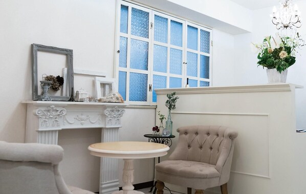 Une pièce avec une cheminée blanche, une table ronde, un fauteuil à capitons, un miroir et une fenêtre avec du verre teinté en bleu.