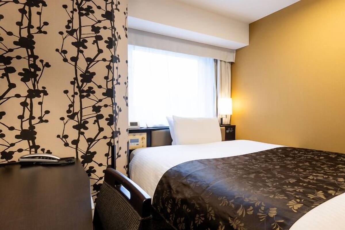 A hotel room with a bed, bedside table, lamp, and a window with curtains.