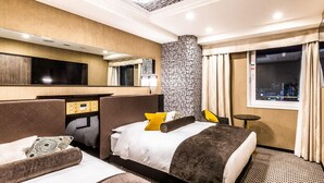 Ein Hotelzimmer mit zwei Betten, einem Schreibtisch, einem Spiegel, einem Fenster mit Stadtblick und einem gemusterten Teppich.