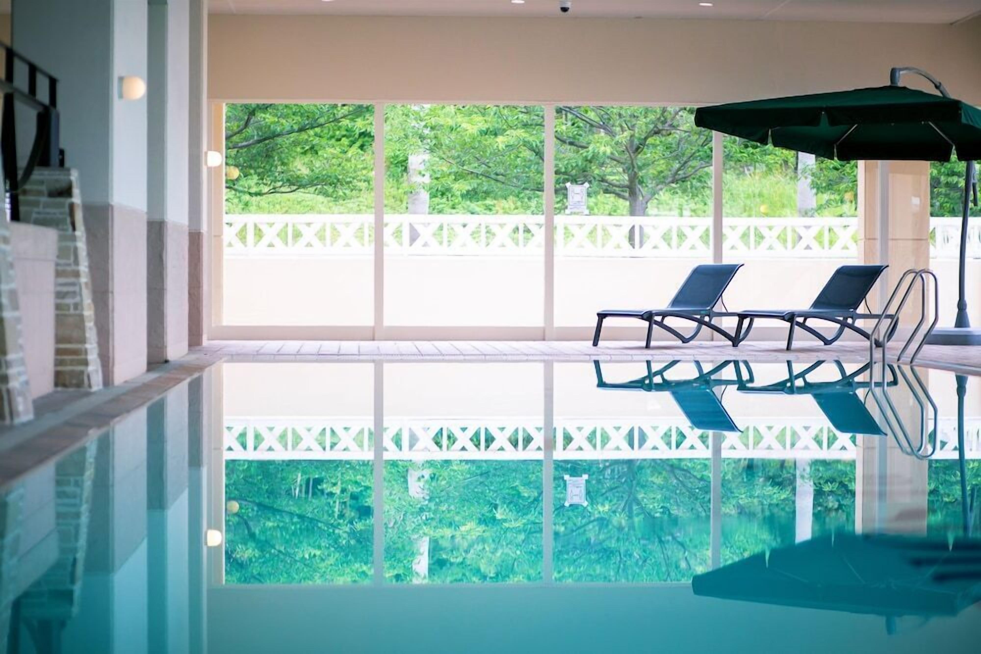 Une piscine intérieure offrant une vue dégagée sur la verdure environnante. On y trouve des chaises longues et un parasol.
