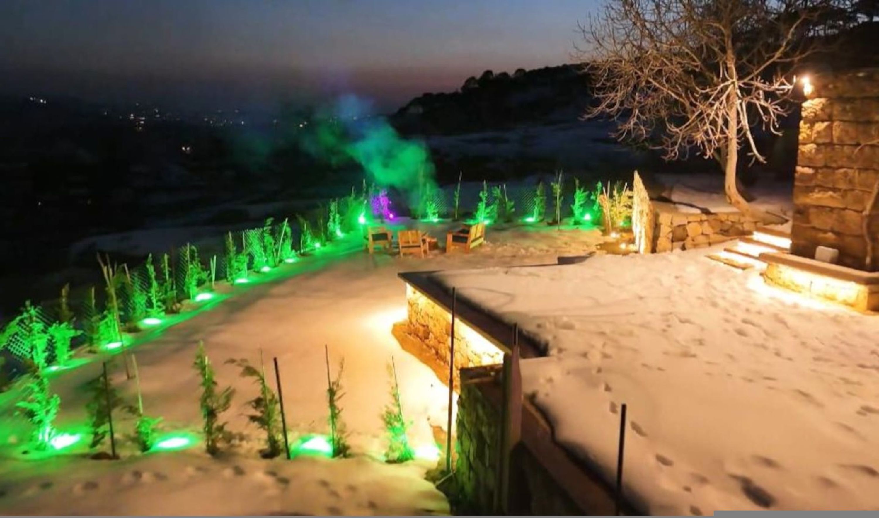 A snow-covered outdoor area with a stone structure and green-lit trees.