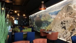 A restaurant interior with a mountain mural, green plants, and orange and blue seating.