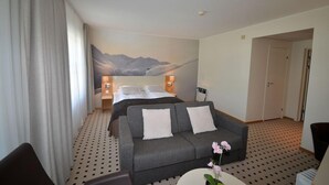 A hotel room with a bed, a grey sofa, a small table with a vase, and a wall mural of mountains.