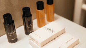 Un tocador de baño con encimera blanca, una botella negra de champú, una botella negra de acondicionador y dos cajas blancas con la etiqueta 'beON Hotel Collection' y 'Gorro de ducha'.
