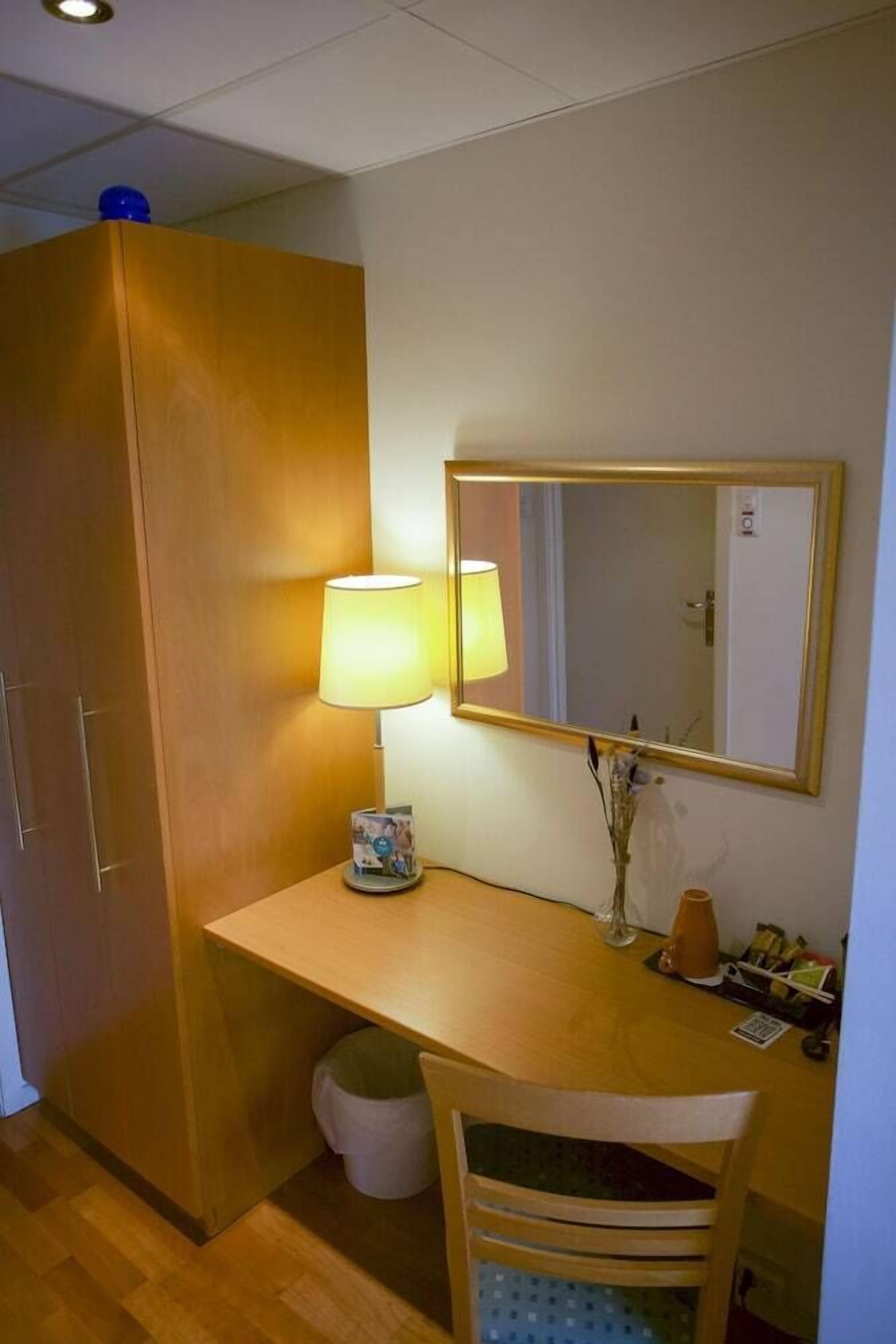 A compact room with a wooden desk, a chair, a lamp, a mirror, and a wardrobe.