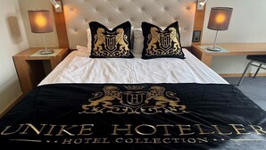A hotel room with a tufted headboard, two bedside lamps, a bed with white and black bedding, and a black throw blanket with gold lettering.