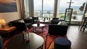 A modern hotel room with a large window offering a scenic view, a cozy seating area with sofas and a coffee table, and a small side table with a lamp.
