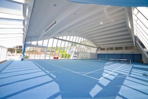 An indoor tennis court with a blue surface, white ceiling, and large windows.