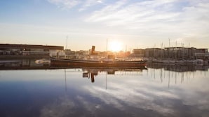 A marina with boats docked, calm water, and a cityscape in the background. - Les Gens de Mer (Dunkirk)