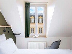A bedroom with a bed, a chair, a window with curtains, a radiator, and a wall-mounted shelf with headphones. - Grand Hotel De Metz (Metz)
