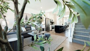 En moderne stue med sofa, sofabord og skammel, et tekøkken med hårde hvidevarer, og en trappe op til et andet niveau.