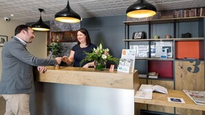Un comptoir d’accueil en bois, une réceptionniste et un client. Au-dessus, on aperçoit des lampes suspendues noires, un mur en briques orné de l’enseigne « Brit Hotel », ainsi que des étagères remplies de divers objets et une horloge.