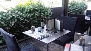 Un espace de restauration en plein air avec une table dressée pour deux personnes, comprenant un menu, une bouteille de vin, des verres et des couverts.