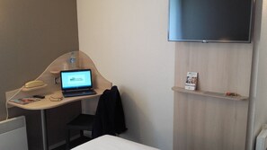 Een hotelkamer met een bureau, een laptop waarop 'BRIT HOTEL' staat, een telefoon, een aan de muur bevestigde televisie en een klein plankje met brochures.