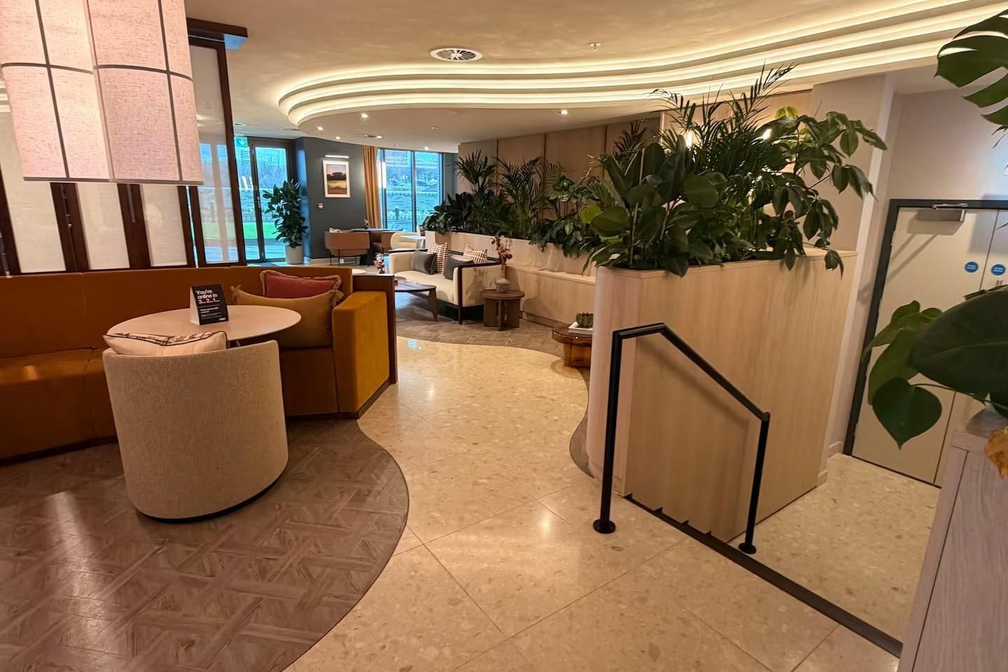 A hotel lobby with seating areas, a reception desk, and large potted plants.