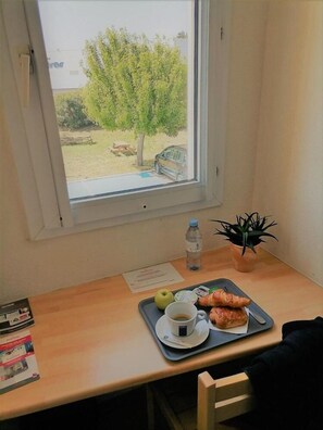 A room with a window, a desk with a tray containing a croissant, a water bottle, and a cup of coffee. - Fasthotel Montpellier ST Jean DE Vedas (Saint-Jean-de-Vedas)