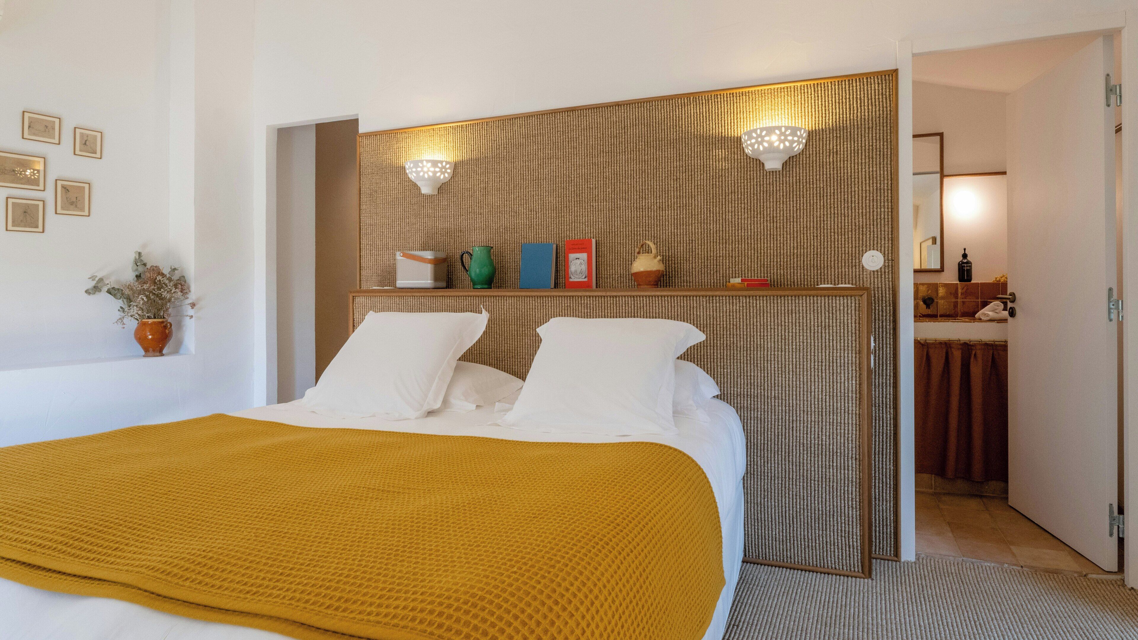 A hotel room with a bed, a bedside table with books and a vase, a wall-mounted light fixture, and a bathroom visible through an open door.