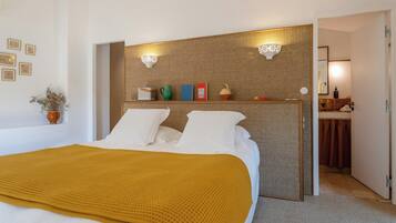 A hotel room with a bed, a bedside table with books and a vase, a wall-mounted light fixture, and a bathroom visible through an open door.