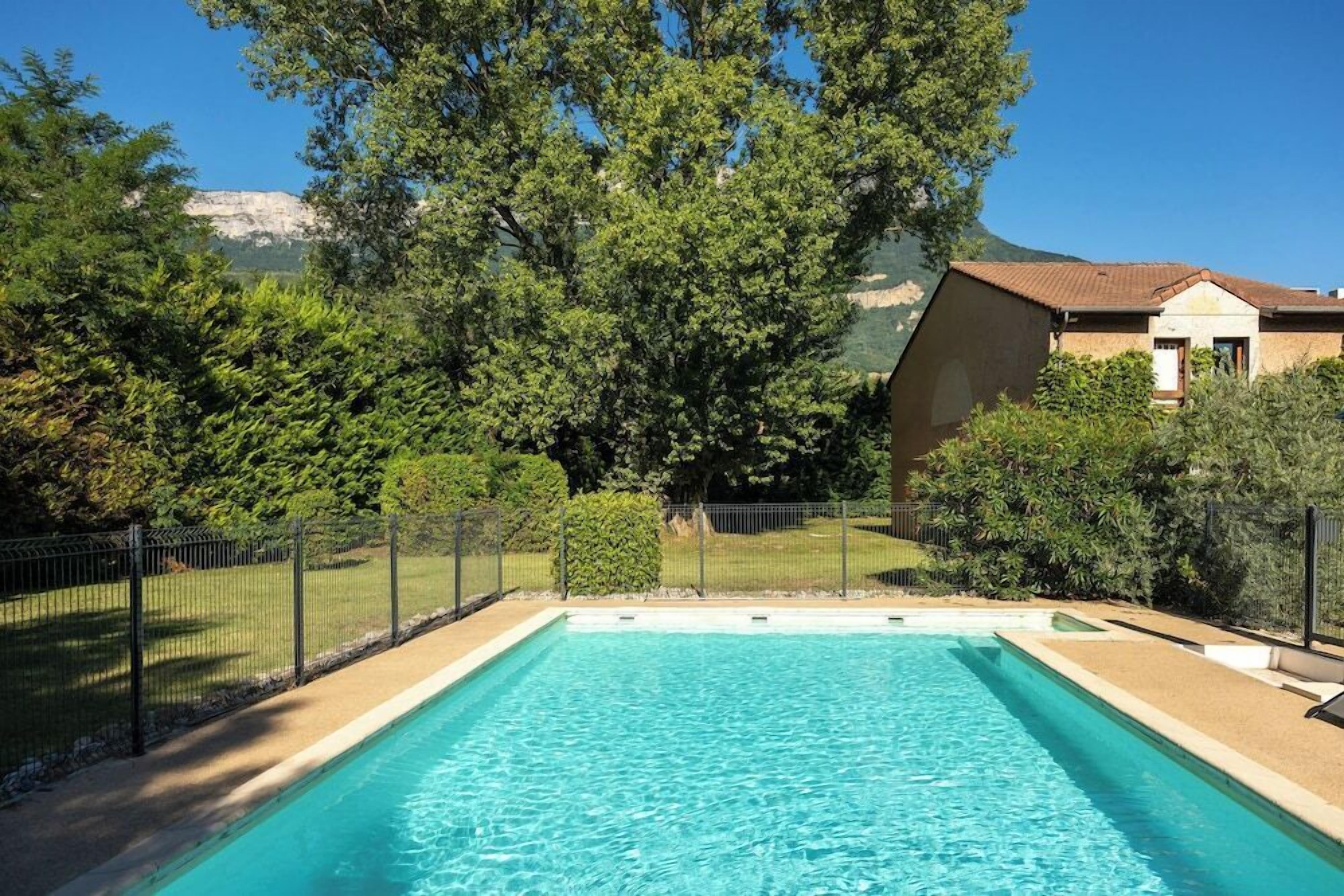 A rectangular swimming pool with clear blue water, surrounded by a fence and greenery, with a building in the background.