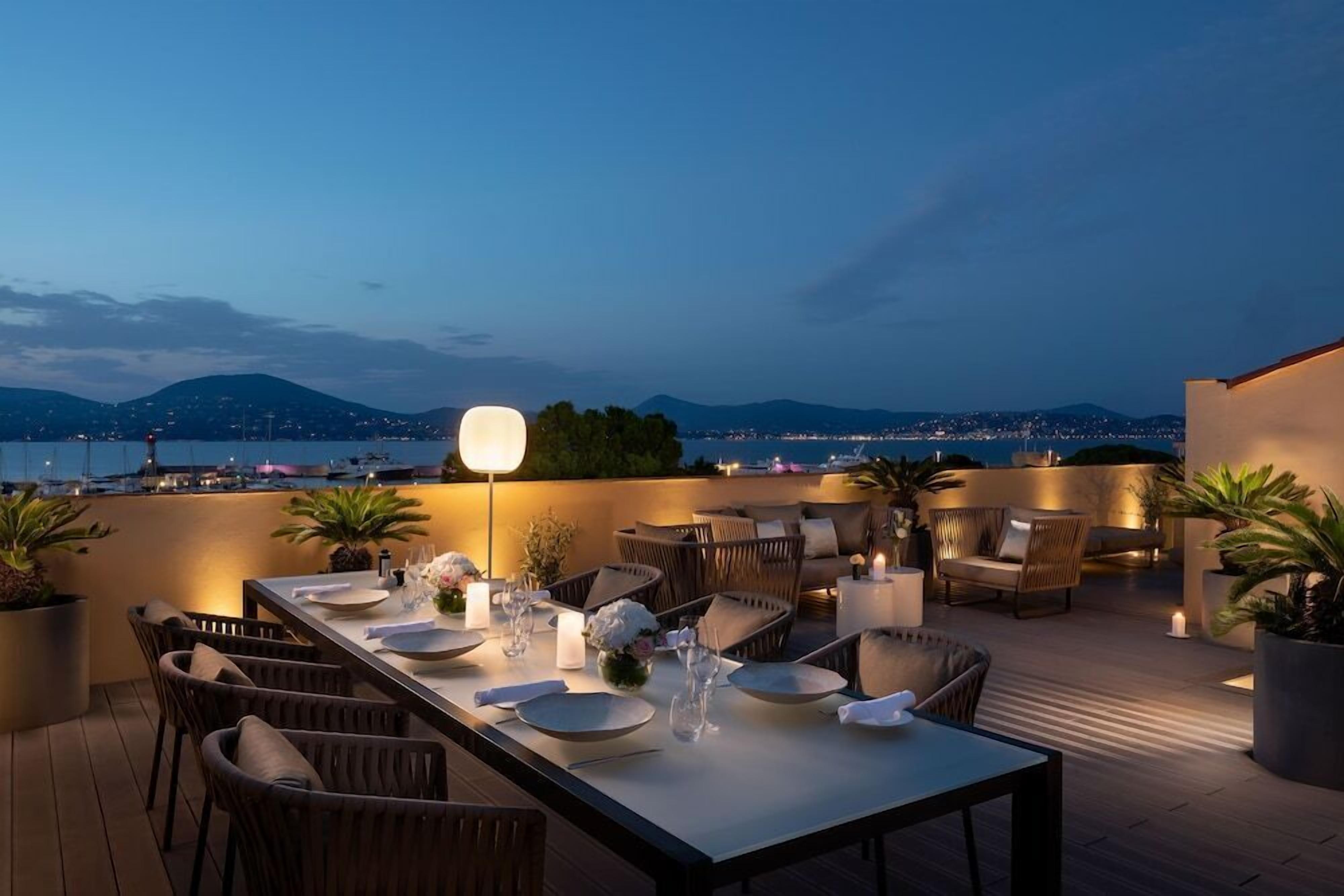 Un área de comedor en la azotea con una mesa preparada para dos, rodeada de sillas de mimbre, plantas en macetas y vistas a la ciudad y montañas al anochecer.