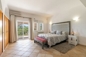 Un dormitorio con cama, mesita de noche, banqueta y vistas al exterior.