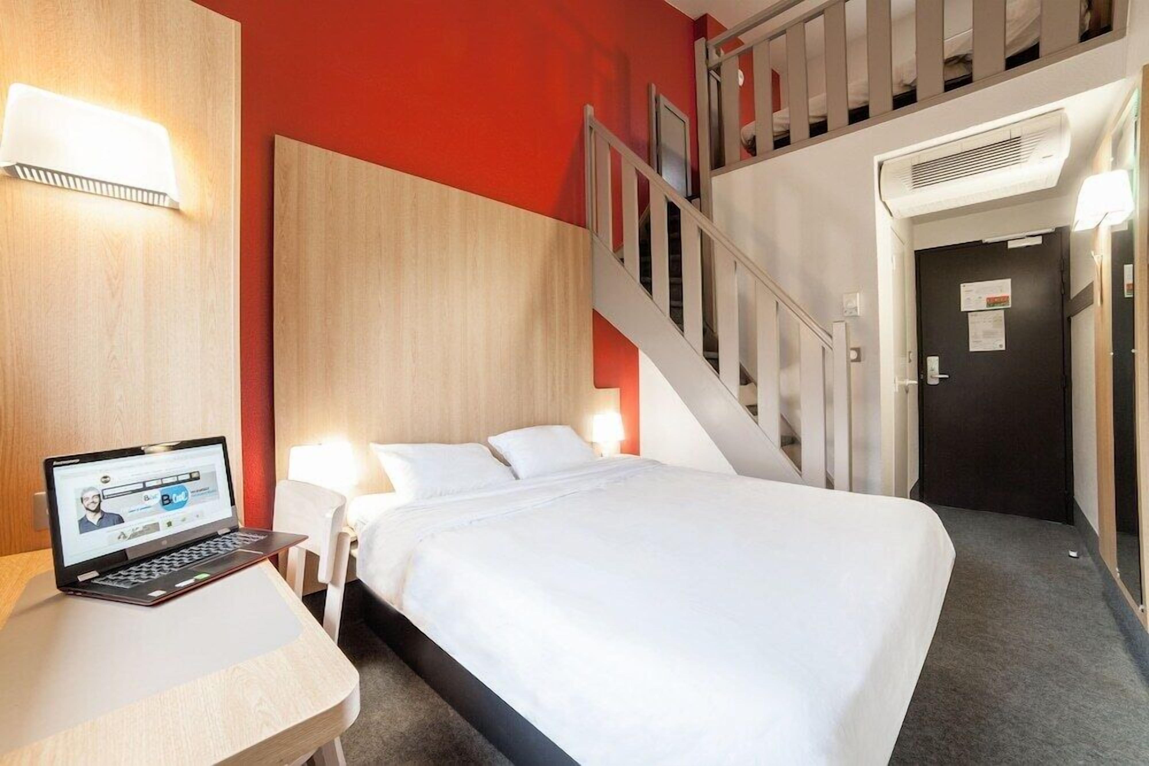 A hotel room with a bed, a desk with a laptop, a staircase, and a door.