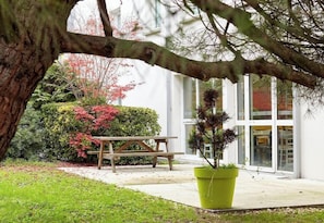 A patio area with a wooden table and chairs, surrounded by greenery and a building with large windows. - B&B Hotel Evry Lisses 3 Etoiles (Lisses)